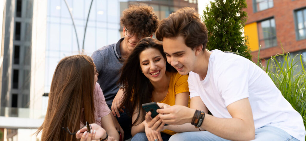 Jovenes reunidos afuera de un edificio sonriendo y viendo un telefono